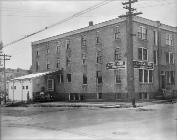 View from across street of a shirtless young man standing beside an automobile in the shade of a lean-to attached to the old Ringling Hotel. Signs on the three-story brick building identify the current occupant as Kingsford Outlet Store and Warehouse. There are many utility lines overhead.
