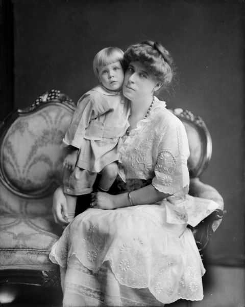 A studio portrait of an unidentified woman seated on a Victorian love seat with a small boy standing beside her. She wears an intricately embroidered dress.