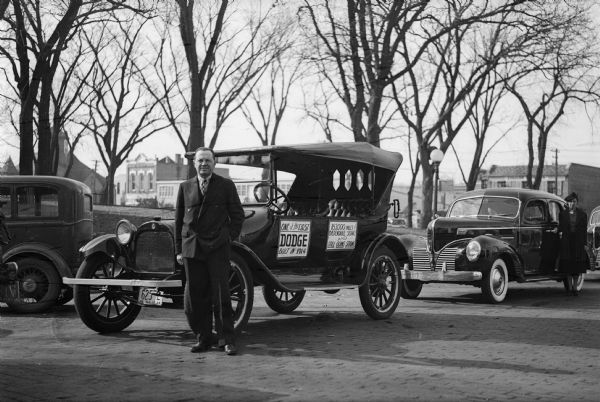 W. L. Tarnutzer stands beside a 1914 Dodge automobile parked on the Sauk County courthouse square. A newer model Dodge with a woman beside it is parked behind the older vehicle. A sign on the bumper identifies it as a "Luxury Liner". The First United Methodist Church is visible in the left background.