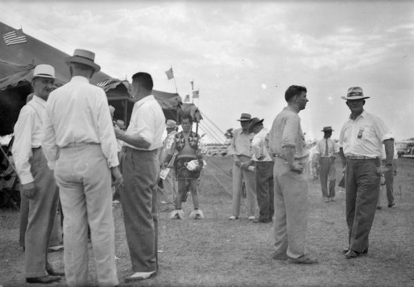 Men in summer clothes stand in small groups outside of a large canvas tent. The men on the right wear badges with ribbons. In the background, a man in Native American dress holds leaflets. His lightning pattern, finger-woven sash is typical of Ho-Chunk hand work.