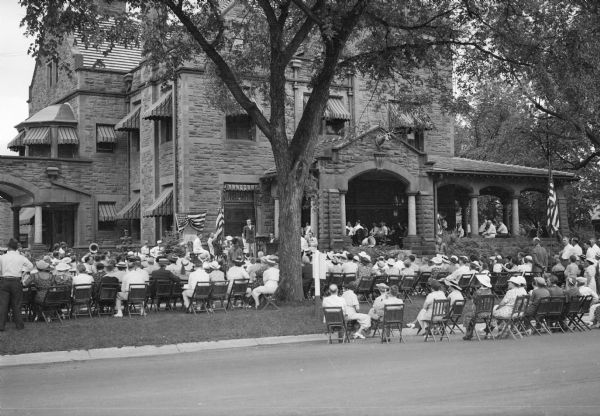 View from across street of a crowd sitting on the porch, lawn and street in front of the Elks Lodge (Al. Ringling house) listening to a speaker standing at a microphone. There are flags and a band near the speaker's platform. Awnings shade the windows of the three-story stone mansion.