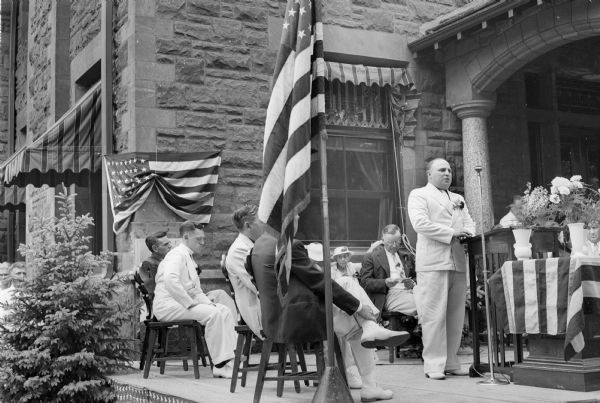 A stocky man in a white suit stands at a lectern and microphone in front of the Elks Lodge (Al. Ringling home). Other men and women sit on the platform. A table covered with an American flag holds vases of flowers.