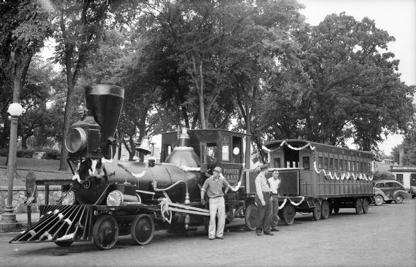 Two men and a police officer stand beside the "Pioneer," an early Chicago and Northwestern Railroad locomotive which is parked beside the Sauk County Courthouse Square. There is a passenger car with a platform attached to the engine and both are draped with garland. There are automobiles and a Cities Service gas station in the background.
