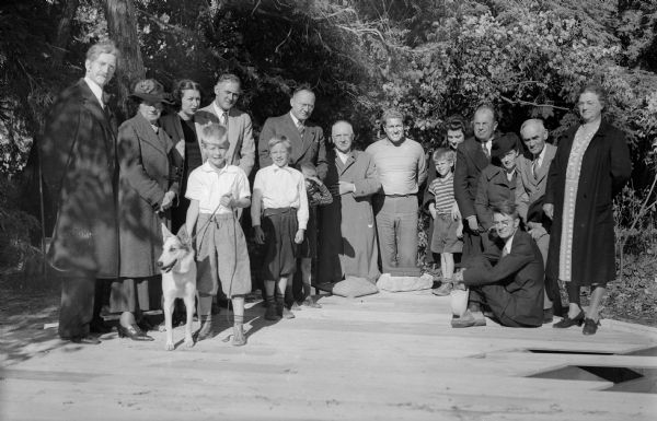 A group poses at the laying of the cornerstone for August Derleth's new house, the Place of Hawks. Attendees include, back row, left to right, E.B. Trimpey; Alice Kent Trimpey; Mary Waterstreet Dessureau; Theodore Brown; Robert Dessureau; Leo J. Weissenborn, architect; August Derleth; Mrs. Harold Bachhuber; Raymond Leuth; Mrs. Charles Brown; Charles E. Brown; and Mrs. George Lins. Front row: Jack Bachhuber (holding dog); Billy Meicher; Richard Bachhuber (with hand shading eyes); David Bachhuber; and, seated, Dr. Harold Bachhuber.