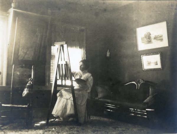 Alice Kent Trimpey sits painting at an easel in front of a window in the Trimpey home. There is a sofa behind her.