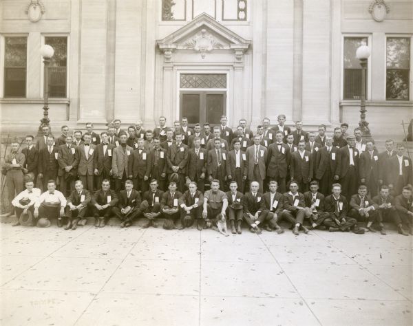 A large group of young men pose on the steps of the Sauk County Courthouse. They would leave that day for the newly constructed Camp Grant near Rockford, Illinois. A dog rests in the center foreground. Most of the men are wearing tags attached to the left lapel on their suit jacket.