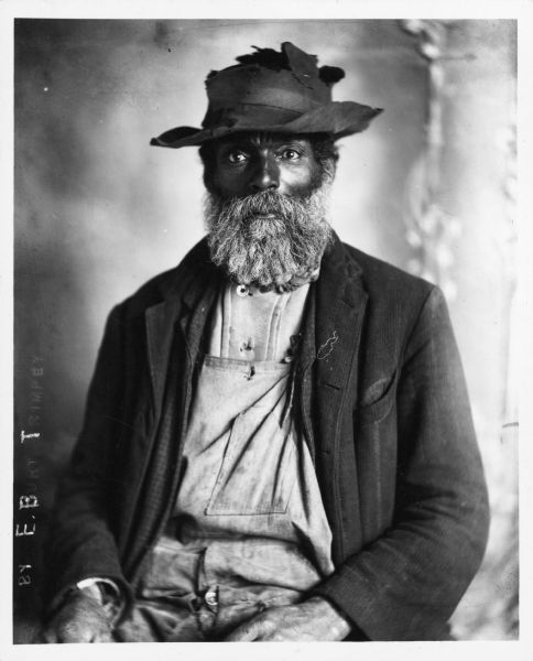A copy print of a studio portrait of a seated African American man with a full beard. He wears a tattered hat and soiled overalls and jacket.