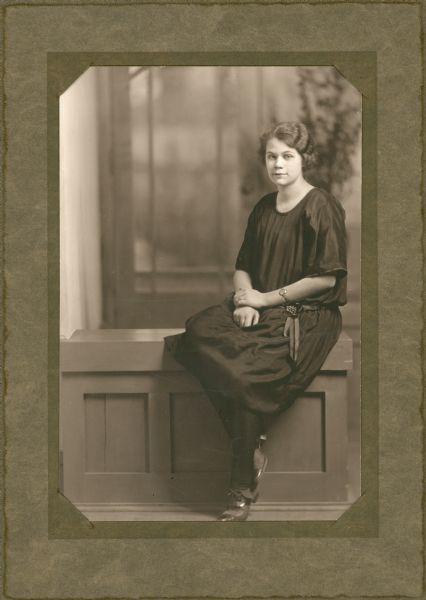 Full-length portrait of a young woman sitting on a half wall inside the photographer's studio. She is wearing a fashionable dress with a ribbon belt, dark stockings and pumps. The rear door of the photographer's studio forms part of the background.