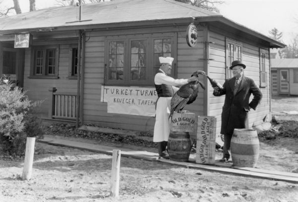 A man wearing a cap and apron holds a (likely taxidermied) Tom turkey while a second man offers the turkey a drink from a bottle. Wooden boxes labelled "Old Gold Lager" are displayed with two barrels on a plank walkway. There is a hand made sign on the small wood frame building identifying it as "Krueger Tavern." An electric sign for "Old Gold on Tap" hangs on the corner of the building.
