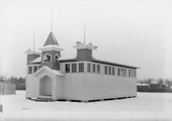 A small, wood frame building with a band of high windows and three short spires stands on the snowy Sauk County Fair Grounds. There is a covered entry porch under the middle spire. Houses are visible beyond a wooden fence in the background.