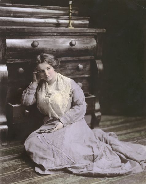 A young woman identified as Mrs. Earl Baltes poses in nineteenth-century attire, seated on the floor with her arm resting on the open bottom drawer of an antique dresser. She is holding a cased photographic image of a person. She is wearing a lace shawl fixed with a brooch.