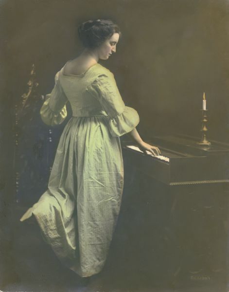 A woman in an old style dress stands in profile by a keyboard instrument with her left knee resting on a Gothic Revival chair. A candle burns in a brass candlestick on the instrument.  The photographer's name, "Trimpey" is embossed in the lower right corner.