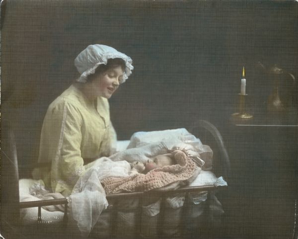 A hand-tinted image of a young woman with a ruffled nightcap tending an infant wrapped in a crocheted blanket lying in a cradle. A candle is burning in a brass candlestick on a table in the background.