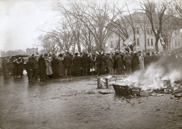 A crowd has gathered facing a corner of the Sauk County Courthouse square where a man, elevated above the crowd, waves an American flag. A bonfire burns on the street in the foreground. Several other American flags are prominently displayed. There is snow on the ground and the trees are bare.