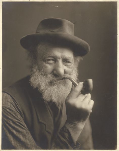 A seated, head and shoulders portrait of a man with a full, gray beard moustache holding a pipe to his mouth. He is wearing a hat and a vest.
