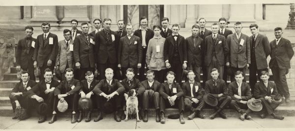 A group of 32 draftees pose on the steps of the Sauk County Courthouse before leaving Baraboo for Columbus Barracks in Ohio. There are paper tags attached to their clothing. A spotted dog sits on the ground in front of the group.