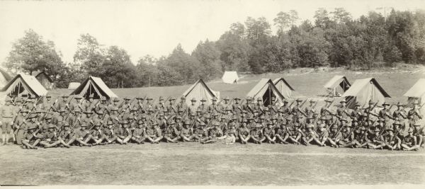 Soldiers of Company I, Baraboo, Wisconsin, pose with their rifles at Camp Grant near Rockford, Illinois. There are tents behind them and a wooded area in the background.