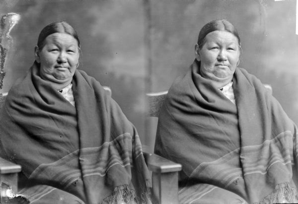 Unidentified Native American woman, probably Ho Chunk, posing for a seated, waist-up stereograph portrait in front of a painted backdrop. She is wearing a fringed blanket around her shoulders.