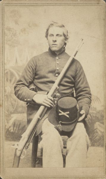 Seated carte-de-visite portrait in front of a painted backdrop of John Crossman, Company C, 1st Wisconsin Heavy Artillery, sitting with a musket across his chest and his hat on his lap.