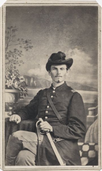 Seated carte-de-visite studio portrait in front of a painted backdrop of 2nd Lieutenant Milo Suley, Company F, 4th Wisconsin Cavalry, sitting in a chair and holding his sword in his left hand.