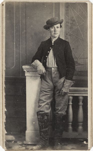 Full-length carte-de-visite portrait in front of a painted backdrop of Private Monroe J. Weatherwax, Company I, 4th Wisconsin Cavalry. He is wearing a hat, gloves, and boots, and is standing in front of a railing, resting his arm on a post.