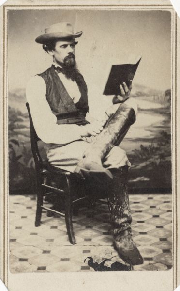 Seated carte-de-visite portrait of James Williams, Company A, 4th Wisconsin Cavalry, sitting and reading a book in front of a painted backdrop.