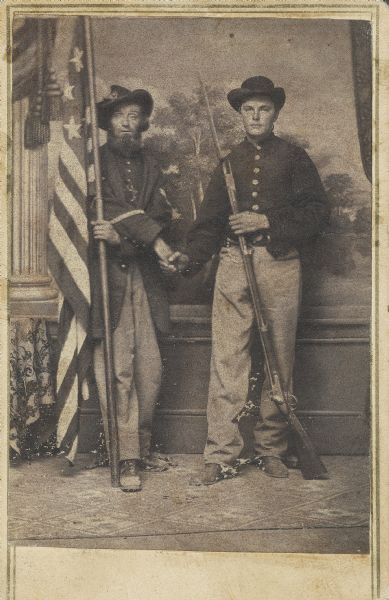 Full-length carte-de-visite of Martin Shenk, holding a flag, and Ryndert Van Loon, holding a musket with bayonet attached. They are posed shaking hands in front of a painted backdrop. Both men were from Company I, 8th Wisconsin Infantry.