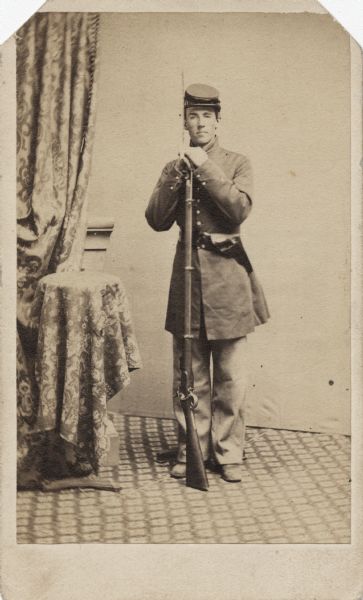 Full-length carte-de-visite studio portrait in front of a backdrop of Corporal Dwight S. Allen, Company C, 22nd Wisconsin Infantry, in uniform, leaning on his rifle.