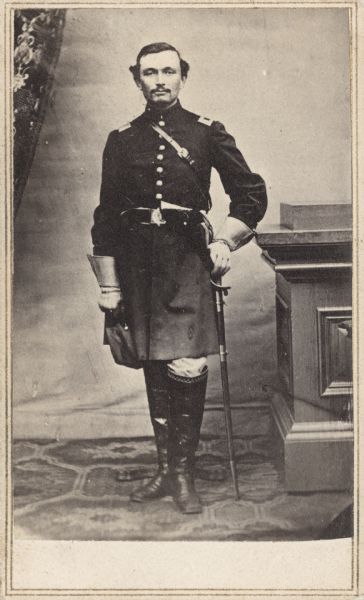 Full-length carte-de-visite portrait of Lieutenant Carlos W. Wellman (or Willman), Companies A and K, 37th Massachusetts Infantry, standing in full uniform grasping his sword. When he was mustered into service he held the rank of 2nd Lieutenant, and he was mustered out of service as a captain.