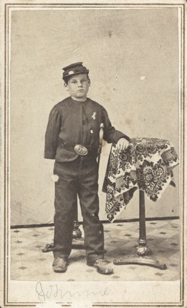 Full-length carte-de-visite portrait of Johnnie Potter standing in uniform. He is standing next to a table, and there is a sword on his left side. The boy is missing his right arm.