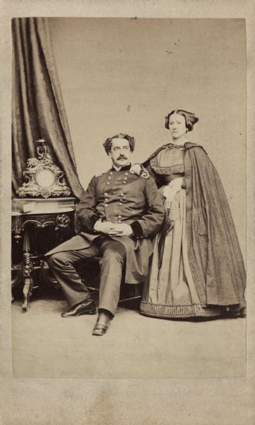 Full-length carte-de-visite of Major General Abner Doubleday in uniform, sitting, and his wife Mary, standing, wearing a cape over her dress.