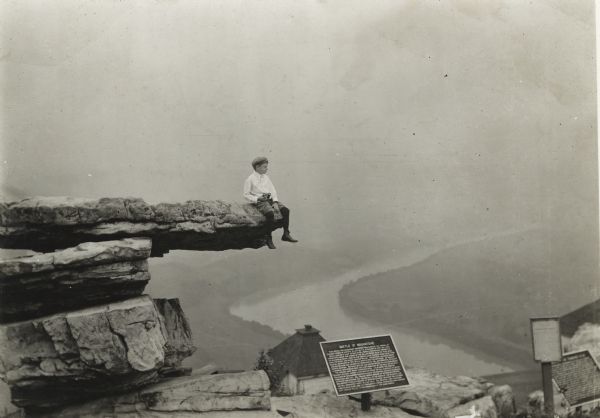 A small boy holding binoculars is sitting on the edge of Umbrella Rock, with a river far below. Below him is a plaque commemorating the Battle of Wauhatchie.