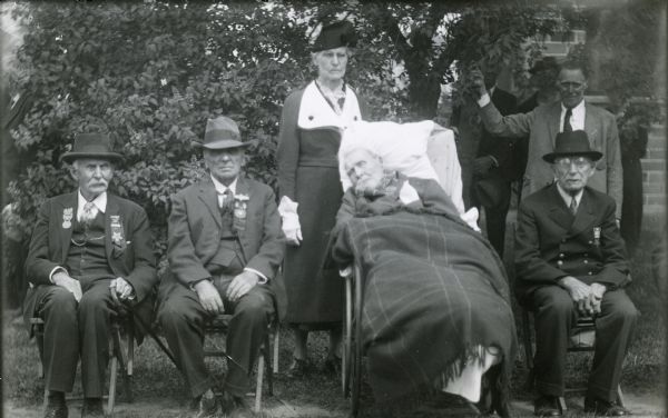 Outdoor group portrait of members of the Grand Army of the Republic; left to right: L.A. Wilcox, Ansel Gailsburg, Elmer Sorkness (in wheelchair), and B. Regli. The other people are unidentified.