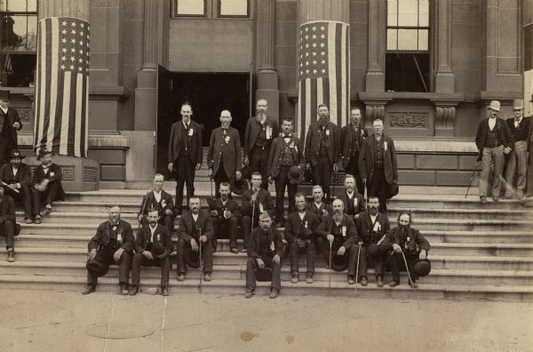 Group portrait outdoors of the Veterans of Company I, 29th Wisconsin Volunteer Infantry at a Grand Army of the Republic reunion. Center group, standing, from left to right: F. Leonard, J. Gould, F.F. French, P. Tubbs, E. Allen, J. H. Welsh, C. Merril. Seated, from left to right: O. Chapman, J. Rector, D. Rice, W. Coon, T. Taylor, J. Kern, M. Andrews, I.M. Allen, R. Alton, J.C. Sayler, T. Stephens, O. Peterson, and J. Gardner. First Captain of the Company, Oliver C. Bissell, was not present.