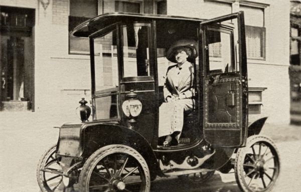 Mrs. Anna Woodward, Kate Quinney's employer, seated in an electric car parked in front of a city building.