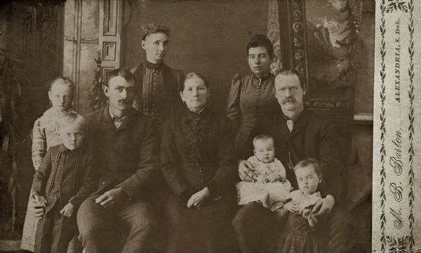 Posed studio portrait in front of a painted backdrop of the Quinney family, including Bridget (O'Keefe) Quinney, her sons Thomas (Tom) and William (Bill), their wives, Florence and Agnes, and four grandchildren.
For an image of Bridget (O'Keefe) Quinney only, see Image ID: 101532.