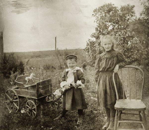 Floyd and Marjorie Quinney playing in the yard at the Old Place.  Floyd has a small wagon and Marjorie stands by a chair.  Possibly taken by a traveling photographer (?).