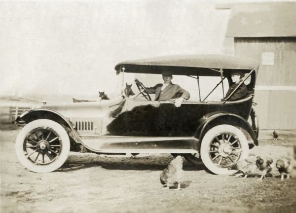 Will and Lorena Holloway seated in a car parked in a farmyard while chickens forage in the foreground.
