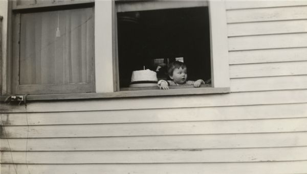 Richard Quinney looks out the window of the farmhouse on his first birthday.  Next to him on the windowsill sits a cake with one candle.
