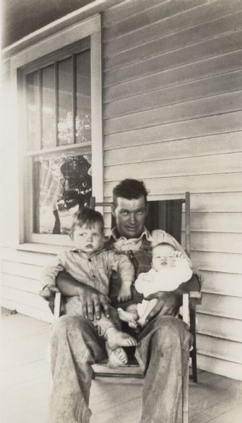 Young Richard Quinney and his new brother Ralph seated on their father's lap in a chair on the farmhouse's porch.