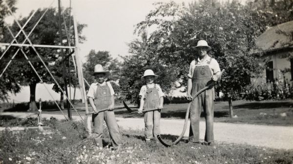 Richard and Ralph Quinney, posing for a photo with their father, Floyd Quinney with corn knives and a scythe.