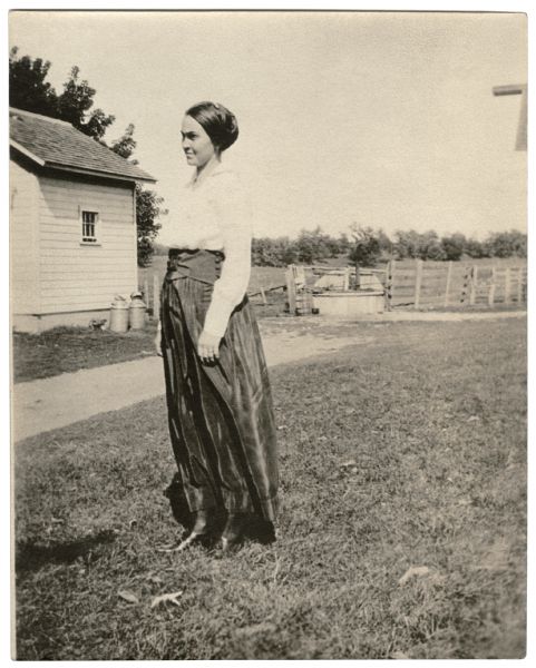 Lorena Taylor Holloway, Richard Quinney's maternal grandmother, standing in a farmyard with a building, milkcans and a fenced field in the background.