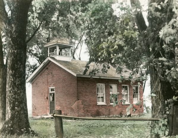Hand-colored photograph of Dunham School, standing in a grove of trees, surrounded by a fence. The building is built of brick and has a bell tower, with a front door with a transom window in the center. The  bricks on the corner have been repaired. Four windows are on the side.