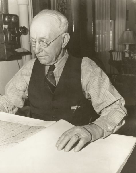 W.V.B. Holloway, Richard Quinney's maternal grandfather, sitting at a a table looking at a large book of maps.  A phone is mounted to the wall next to him and in the background a living room scene is visible.