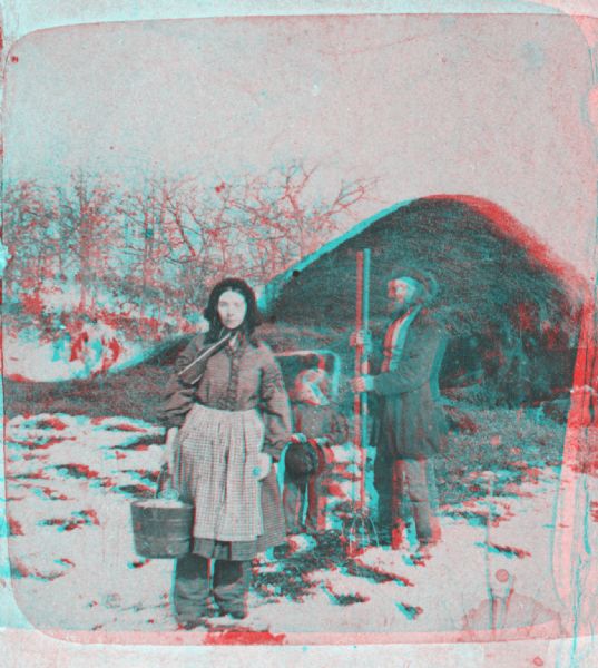 Stereograph of woman, man and child outdoors on a farm with snow on the ground. The woman is wearing bloomers and is holding a bucket in the process of doing chores, the man is holding a pitchfork, and the child is holding a hat. In the background are cows eating near a large pile of hay.