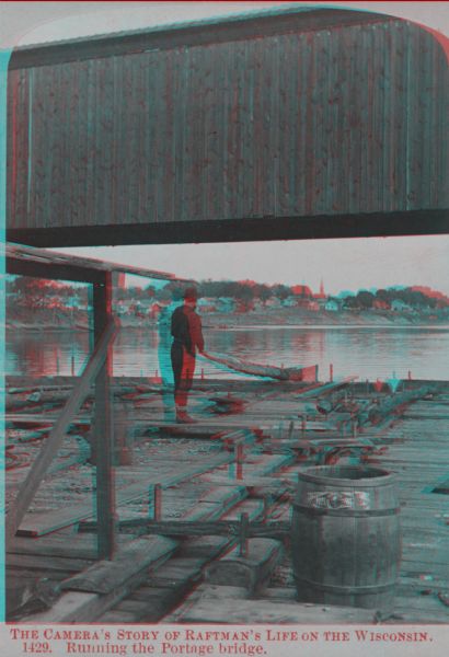 Stereograph of a man standing on the back of a raft holding a hand-hewned oar. The raft is passing under a wooden bridge. A town, including a church, are in the background.