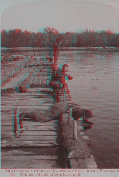 Stereograph of a man lying on his stomach on a raft drinking water from the river. A second man is crouching on the edge of the raft, while a third man is standing lifting something to his mouth. In the distance is a raft at the shore.