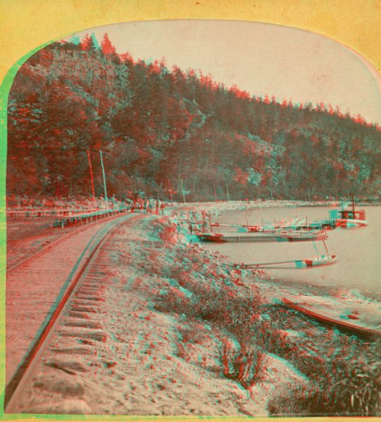 A yellow mount stereograph of the railroad tracks skirting the edge of Devil's Lake, with several boats tied up alongside. From the stereograph series "The Wonders & Beauties of Nature, Devil's Lake, Wis."