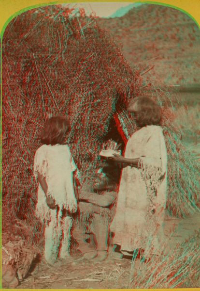 Stereograph image of a Native American man and woman with a young child, members of the Ute Indian Tribe, standing in front of a grass structure.