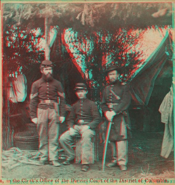 Stereograph of three U.S. Army soldiers in front of tents in camp under a framework of branches.